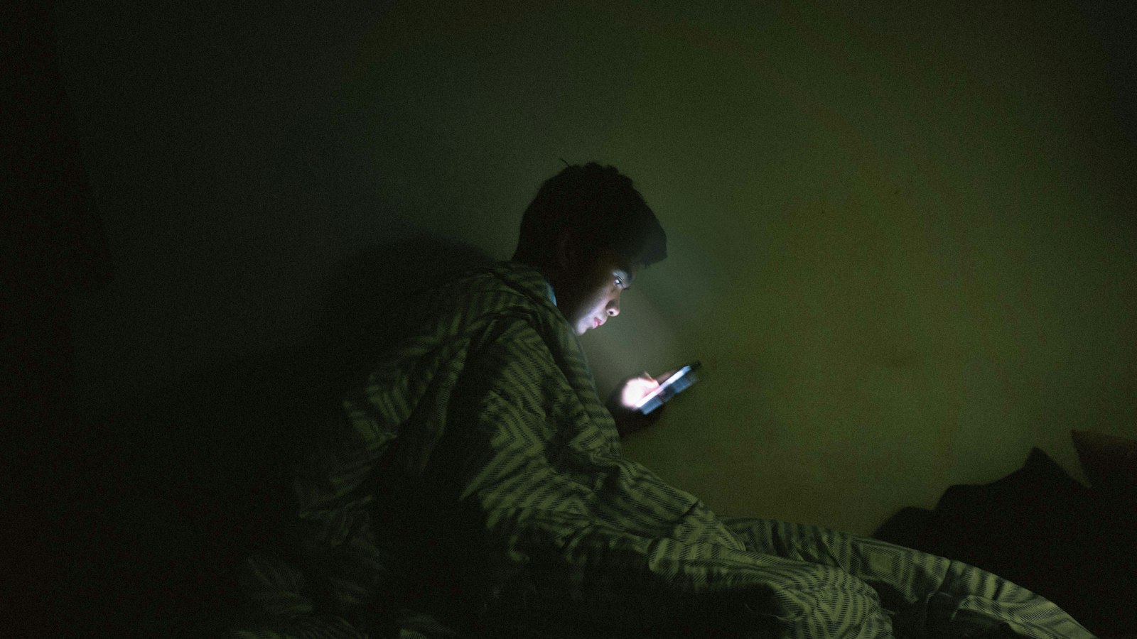 Gece battaniye altında telefonda doomscrolling yaparak olumsuz haberleri kaydıran bir adam