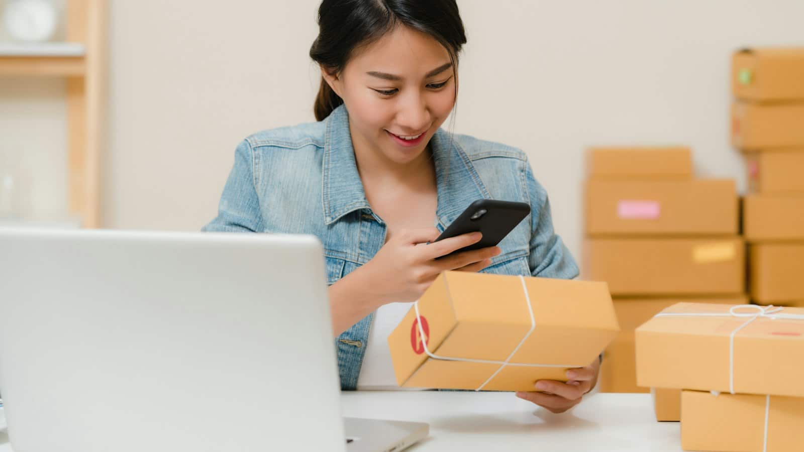 E-ihracat siparişlerini dizüstü bilgisayar ve kargo kutuları arasında telefondan kontrol eden bir kadın